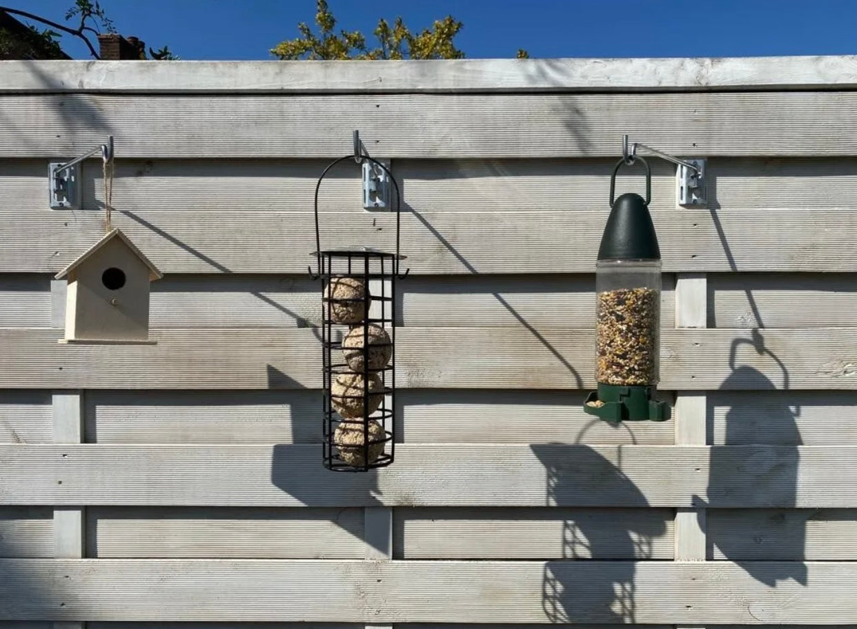 Image shows a white garden fence with 3 bird feeder hanger bars with brackets fitted horizontally holding a bird house, a seed bird feeder, and a fat ball bird feeder. Hanger bar has a deep hooked end to securely retain a bird feeder, etc. Purchased in a 3 pack for £6.99 from potmagic.co.uk. Can be quickly screw fitted to fences, pergolas and walls with screw fittings provided. Brackets are made from strong galvanised steel. Hook-ended hanger bar is made from strong 6mm diameter shiny galvanised steel.